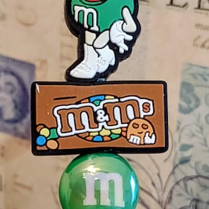 May include: A green M&M's themed pen featuring a green M&M's character, the M&M's logo, and a green ball with a white "m". The pen has a green top and a decorative base.