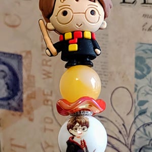 May include: A decorative pen featuring a cartoon character design. The pen has a red cap and a series of decorative beads, including a character with glasses, a yellow bead, and a red floral accent. The pen is designed with a red and silver base.
