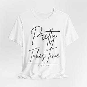 Può includere: T-shirt bianca con la scritta "Pretty Takes Time" in un elegante carattere corsivo. Sotto, la parola "DARLIN'" è visualizzata con un piccolo simbolo a cuore. La maglietta ha un girocollo e maniche corte.
