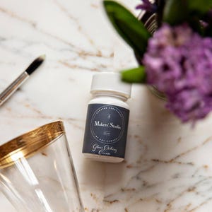 May include: A bottle of Makers' Studio Glass Etching Cream sits on a marble surface, next to a paintbrush and a gold-rimmed glass. The label is dark blue with white text. Purple flowers are in the background.