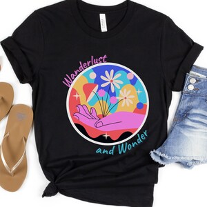 May include: Black t-shirt with a colorful graphic of a hand holding flowers in a circle. The text "Wanderlust and Wonder" is written below the graphic.