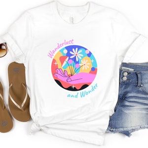 May include: White t-shirt with a colorful graphic design of a hand holding flowers. The graphic is surrounded by a circle with the text "Wanderlust and Wonder".