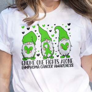 Könnte beinhalten: Weißes T-Shirt mit einem grünen, schwarzen und weißen Gnom-Design. Die Gnome halten grüne Bänder und Herzen. Der Text "Gnome One Fights Alone Lymphoma Cancer Awareness" ist unter den Gnomen gedruckt.