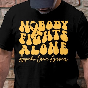 Puede incluir: Camiseta negra con el texto amarillo "NOBODY FIGHTS ALONE", una cinta amarilla y las palabras "Appendix Cancer Awareness". La camiseta la lleva una persona. El diseño está centrado.