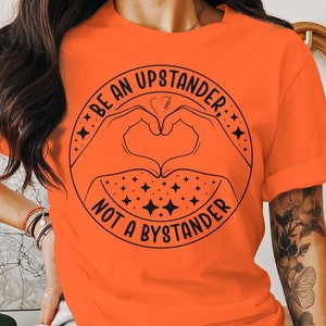 May include: Bright orange t-shirt featuring a black graphic. The graphic includes the text "BE AN UPSTANDER, NOT A BYSTANDER" in a circular design, with hands forming a heart shape in the center. The design also includes stars.