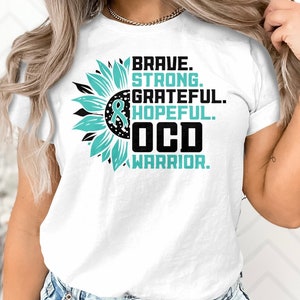 May include: White t-shirt featuring a teal sunflower and the text "BRAVE. STRONG. GRATEFUL. HOPEFUL. OCD WARRIOR."