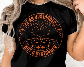 Könnte beinhalten: Schwarzes T-Shirt mit einem orangefarbenen Grafikdesign, auf dem "Sei ein Fürsprecher, kein Zuschauer" steht. Die Grafik zeigt zwei Hände, die eine Herzform mit Sternen drumherum bilden.