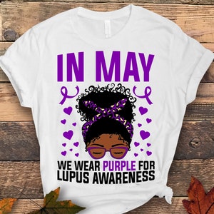 May include: White t-shirt with the text "IN MAY WE WEAR PURPLE FOR LUPUS AWARENESS" in purple. A black woman with a purple headband and glasses is in the center of the text. There are purple ribbons and hearts around the woman.