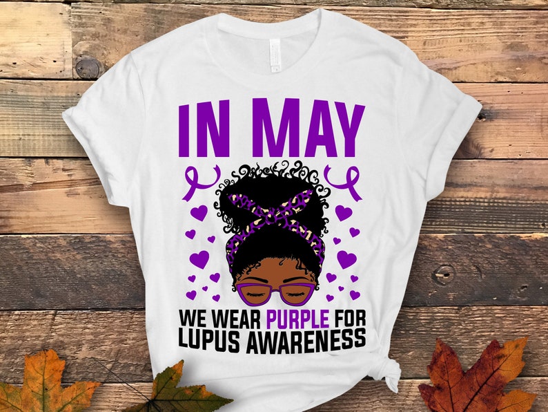 May include: White t-shirt with the text "IN MAY WE WEAR PURPLE FOR LUPUS AWARENESS" in purple. A black woman with a purple headband and glasses is in the center of the text. There are purple ribbons and hearts around the woman.