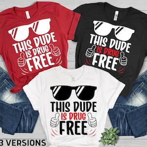 May include: Three t-shirts in red, black, and white, each with the text "THIS DUDE IS DRUG FREE" in a bold, playful font. The design includes sunglasses and a thumbs-up gesture. The shirts are displayed with blue jeans.