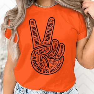 May include: Orange short-sleeved t-shirt with a black peace sign graphic. The peace sign is made up of the words "peace", "love", "unity", "joy", "faith", "hope", "accept", "include", "kindness", and "inclusion".