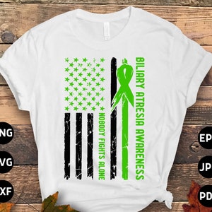 Op de afbeelding: Een wit T-shirt met een groen lint en een Amerikaans vlagontwerp. De tekst "Biliary Atresia Awareness" en "Nobody Fights Alone" staat op het T-shirt gedrukt.