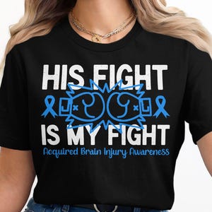 May include: Black t-shirt with white text that reads "His Fight Is My Fight" and "Acquired Brain Injury Awareness". The text is surrounded by blue ribbon symbols and a graphic of two boxing gloves.