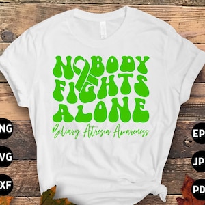 Op de afbeelding: Wit T-shirt met een groene afbeelding met de tekst "Nobody Fights Alone" en een groen lint. De tekst "Biliary Atresia Awareness" staat onder de afbeelding.