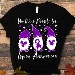 May include: Black t-shirt with a purple, white, and gray design of three gnomes holding purple hearts. The gnomes are wearing purple hats and have white beards. The text "We Wear Purple for Lupus Awareness" is printed on the shirt.