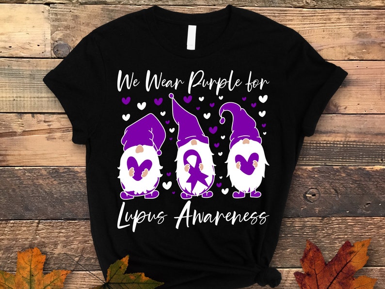 May include: Black t-shirt with a purple, white, and gray design of three gnomes holding purple hearts. The gnomes are wearing purple hats and have white beards. The text "We Wear Purple for Lupus Awareness" is printed on the shirt.