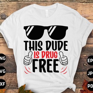 May include: White t-shirt with the text "THIS DUDE IS DRUG FREE" in black and red. The design includes black sunglasses and thumbs-up gestures. The shirt is displayed on a wooden surface.