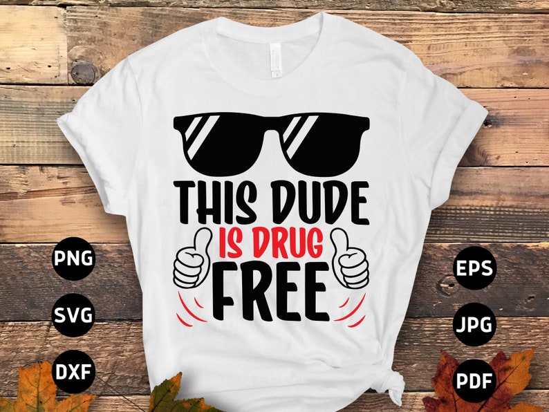 May include: White t-shirt with the text "THIS DUDE IS DRUG FREE" in black and red. The design includes black sunglasses and thumbs-up gestures. The shirt is displayed on a wooden surface.