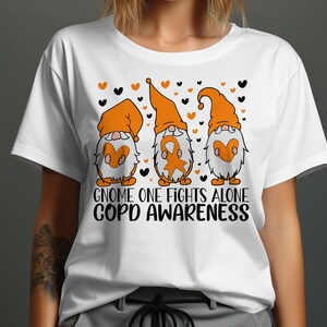 Puede incluir: Camiseta blanca con tres gnomos naranjas que sostienen corazones y cintas naranjas. Los gnomos tienen ojos negros y sombreros negros. El texto "Gnome One Fights Alone COPD Awareness" está impreso debajo de los gnomos.