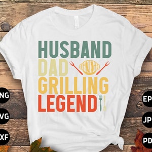 May include: White t-shirt with the words "HUSBAND DAD GRILLING LEGEND" in teal, yellow, and red. The design includes grilling tools. The shirt is a casual wear item.