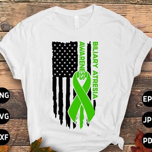 Op de afbeelding: Een wit T-shirt met een verweerd Amerikaans vlagontwerp. Een groen lint met de woorden "Biliary Atresia Awareness" is over de vlag gelegd.