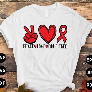 May include: White t-shirt with the words "PEACE LOVE DRUG FREE" in black font. The design includes a peace sign, a heart, and a ribbon, all in red. The shirt is displayed on a wooden surface.