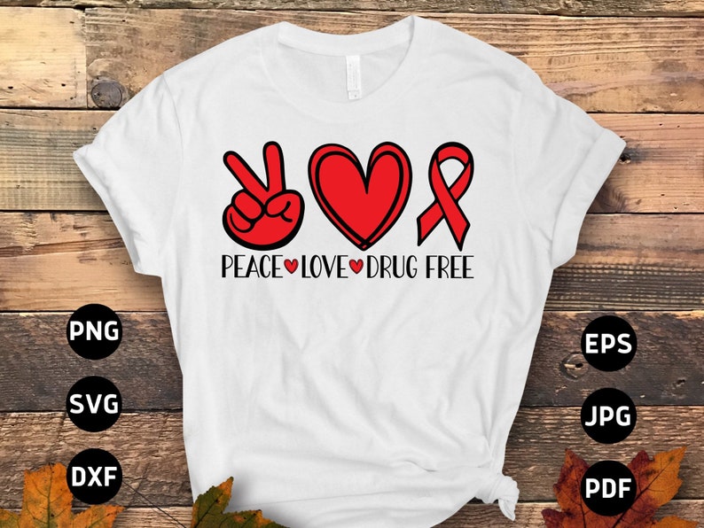 May include: White t-shirt with the words "PEACE LOVE DRUG FREE" in black font. The design includes a peace sign, a heart, and a ribbon, all in red. The shirt is displayed on a wooden surface.