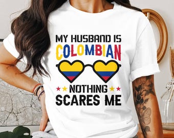 May include: White t-shirt with the text "MY HUSBAND IS COLOMBIAN NOTHING SCARES ME". The word "COLOMBIAN" is in yellow, blue, and red, with heart-shaped sunglasses in the same colors. The shirt is a casual style.