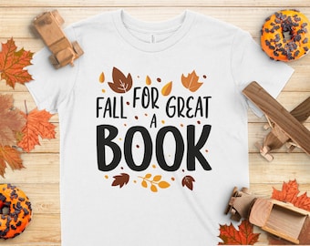 May include: White t-shirt with the text "FALL FOR A GREAT BOOK" in black, surrounded by autumn leaves. The shirt is on a wooden surface with fall-themed decorations, including toy cars and donuts.