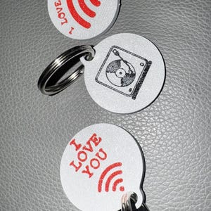 May include: Three white keyrings with silver rings. Two keyrings have the text "I LOVE YOU" in red with a red wifi symbol. The third keyring has a black and white record player graphic.