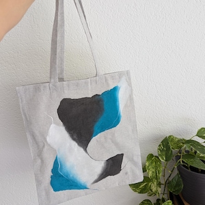 May include: A gray tote bag with abstract art in shades of blue, black, and white. The tote bag is held up by a hand, showcasing the design. The bag has long straps and is made of a light-colored fabric.