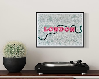 May include: A framed wall art print featuring a detailed map of London with a dark green winding river line and the word LONDON in bold pink 3D-style lettering centered across the map, displayed in a simple black frame.