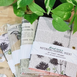May include: A set of hand-sewn notebook journal inserts featuring vintage newspaper print and botanical illustrations. Each insert is wrapped in a white paper band labeled Hand-sewn NOTEBOOK/JOURNAL INSERT, A6, Lined, 100 pages, 28 lb off-white speckled paper.