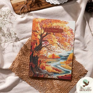 Puede incluir: Un cuaderno con una ilustración con temática otoñal. La portada presenta una representación detallada de un árbol con hojas naranjas y amarillas, un río y el texto "In My Autumn Era". El cuaderno descansa sobre papel marrón y tela blanca.