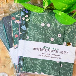 May include: A set of A6 hand-sewn notebook journal inserts with floral and botanical patterns in green, blue, and black. Each notebook features 100 lined pages of 28 lb off-white speckled paper, secured with a gold-tone metal button closure.