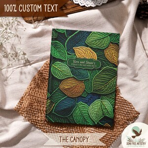 3 of 5 Forest aesthetic journal gifts for hikers. This woodland notebooks is called The Canopy and is displayed on a beige outdoor picnic blanket. A rustic faux embroidered nature journal featuring faux 3d leaf artwork on the cover. It is perfect for family camping memories, hiking adventures and forest lovers or all ages.
