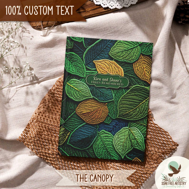 3 of 5 Forest aesthetic journal gifts for hikers. This woodland notebooks is called The Canopy and is displayed on a beige outdoor picnic blanket. A rustic faux embroidered nature journal featuring faux 3d leaf artwork on the cover. It is perfect for family camping memories, hiking adventures and forest lovers or all ages.