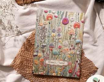 May include: A white journal with a colorful embroidered floral design. The journal cover features the text "Kammy's Journal" in a brown oval frame.