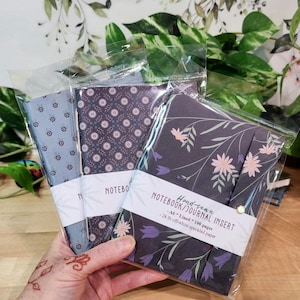 May include: Three hand-sewn A6 notebook journal inserts in clear plastic sleeves. The front notebook features a dark floral print with pink and purple flowers. The middle has a dark geometric pattern, and the left has a light blue bee print. Each has a white paper band with text.