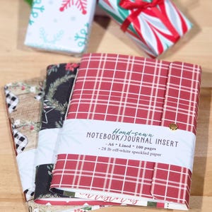 May include: Three hand-sewn notebooks with patterned covers, including red plaid, black with pine branches, and a burlap print. A white paper band on the front reads: Hand-sewn NOTEBOOK/JOURNAL INSERT, A6, Lined, 100 pages, 28 lb off-white speckled paper.