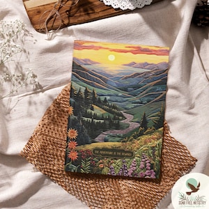 May include: A journal with a landscape painting of a mountain valley at sunset. The artwork features a river, trees, and wildflowers in shades of green, blue, orange, and purple. The text "THE ADVENTURE IS THE REWARD" is at the bottom.