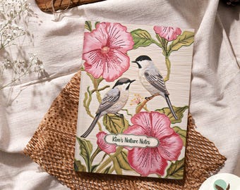 May include: A white journal with a floral design featuring pink hibiscus flowers and two gray birds. The journal has the text "Kim's Nature Notes" on the front cover.