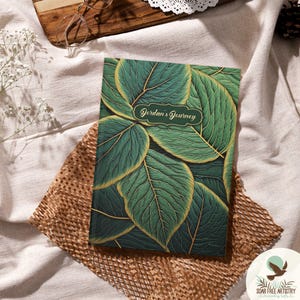 Può includere: Un diario verde con accenti in foglia d'oro e un motivo a foglie. La copertina ha un ovale dorato con il testo "Jordan's Journey" in un carattere corsivo.