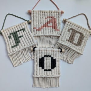 以下が含まれることがあります： 木製の棒が付いたマクラメの壁掛けが4つ。それぞれの壁掛けには、アルファベットの異なる文字「F」、「O」、「D」、「I」が、異なる色で描かれています。「F」は緑、「O」は黒、「D」は茶色、「I」はピンクです。