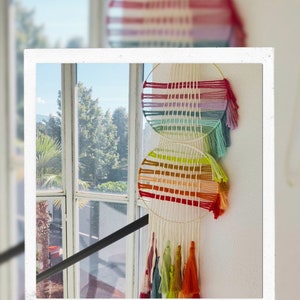 Puede incluir: Un tapiz de macramé con dos diseños circulares tejidos en colores del arcoíris. La pieza presenta borlas de varios colores y el texto "Tapiz de colores" en la parte inferior. El fondo incluye una ventana y un vistazo al exterior.