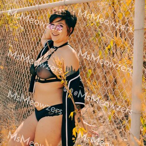 May include: Black lingerie set featuring a sheer bra with harness detailing and matching lace underwear. A black and white jacket is draped over the shoulders. The background includes a chain-link fence and foliage.