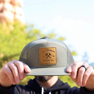 May include: A green and brown trucker hat with a brown leather patch that says "Big Thunder Mining Co." in black lettering. The patch has a crossed pickaxe design.