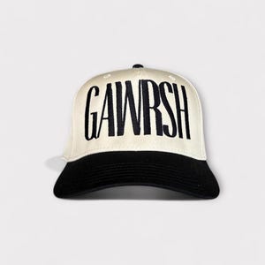 May include: A baseball cap with a cream-coloured crown and a black brim. The word "GAWRSH" is embroidered in bold, black letters across the front of the cap. The cap has a curved brim and a button on top.