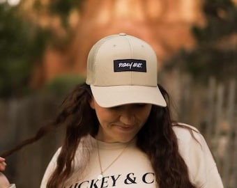 Puede incluir: Una mujer con una sudadera beige con el texto "Mickey & Co. Est. 1928" y una gorra de camionero de malla beige y negra con el texto "Pixley Ave." en negro.