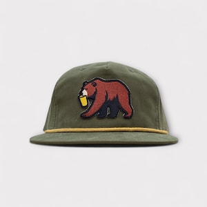 May include: Olive green baseball cap with a brown bear patch holding a yellow cup. The bear is embroidered with black and brown details. The cap has a yellow rope detail along the brim.
